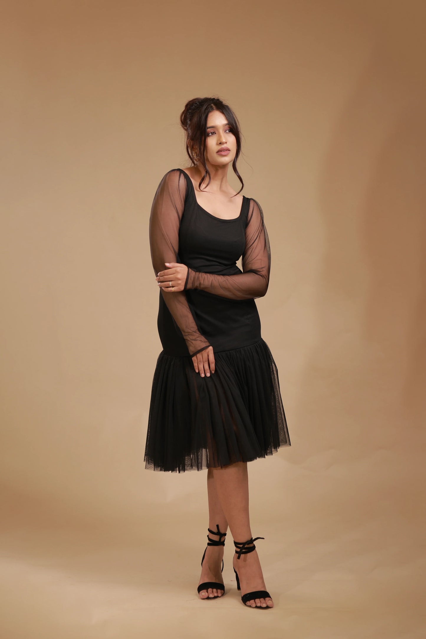Woman in black mesh sleeve dress and heels posing against beige studio background