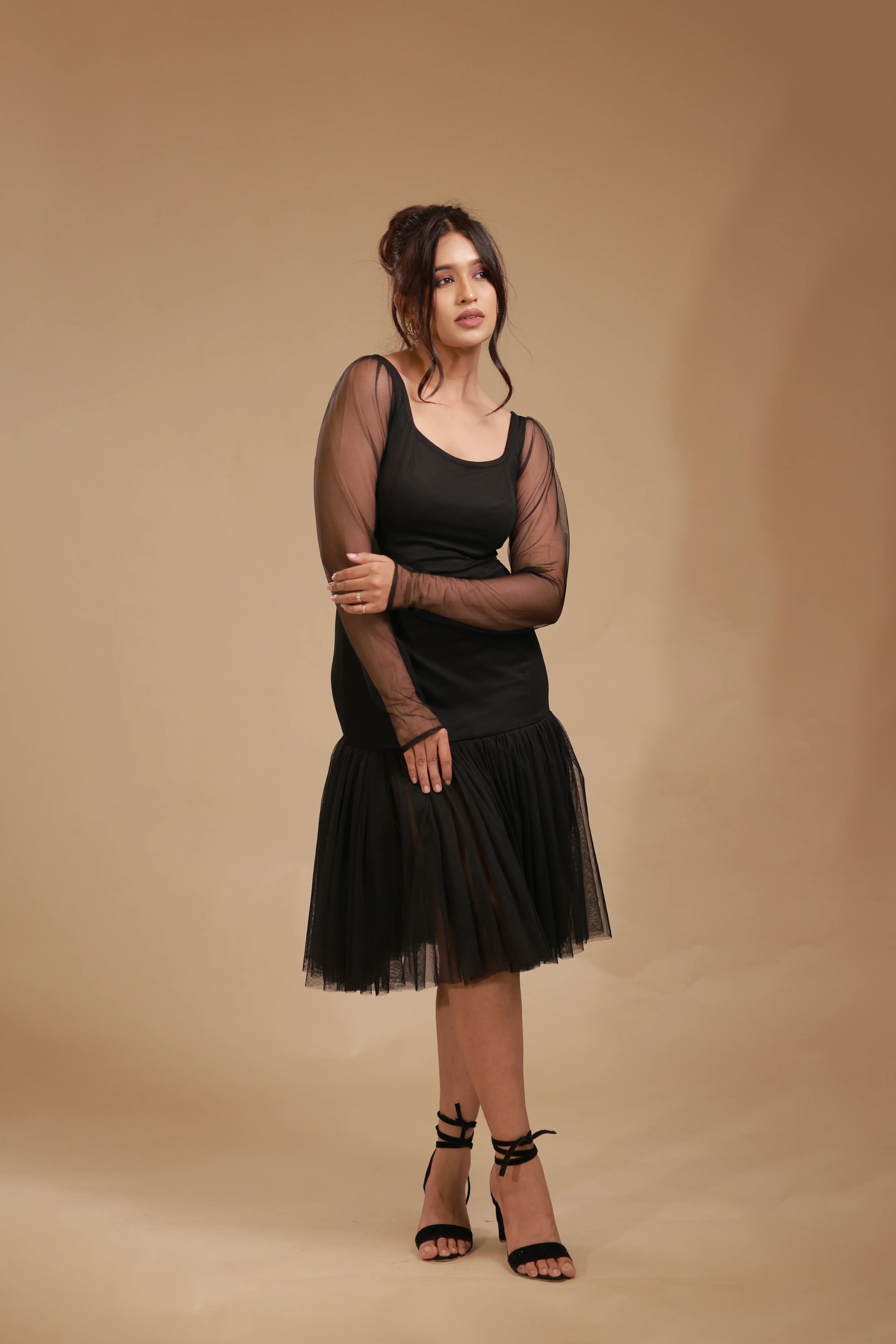 Woman in black mesh sleeve dress and heels posing against beige studio background