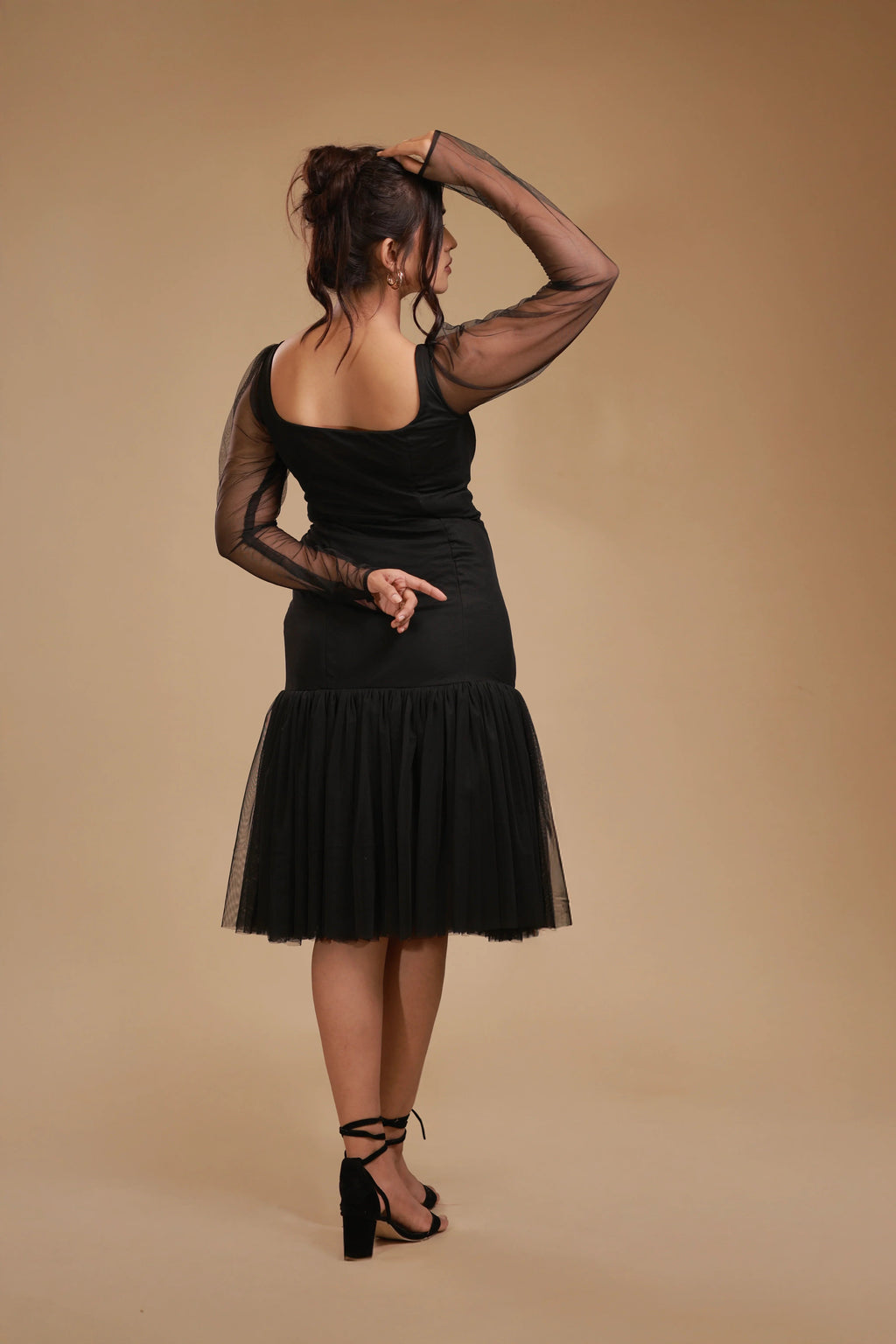 Woman in black mesh sleeve dress with ruffled skirt and block heels, beige background