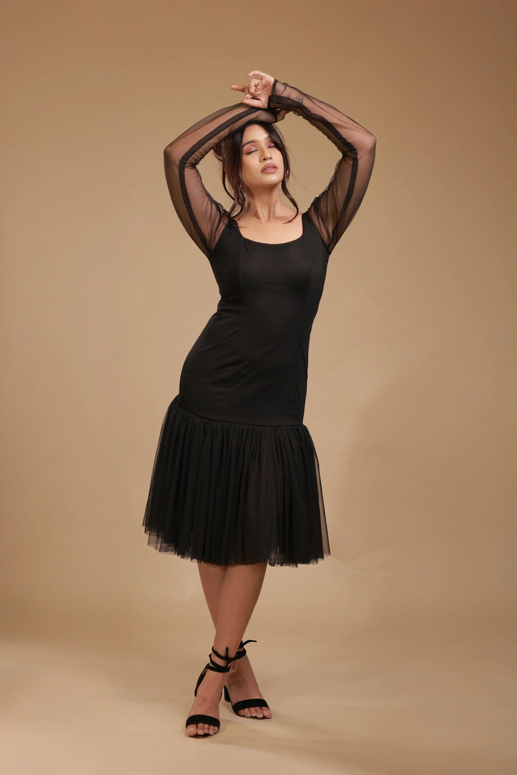 Woman in black mesh long sleeve dress with tulle hem and black heels on beige background